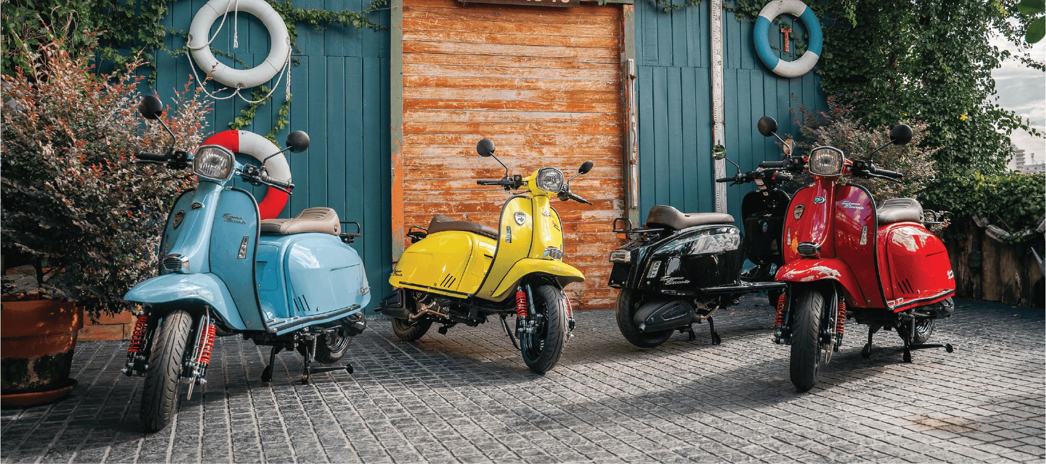 Four Scomadi Turismo Piccolo scooters in blue, yellow, black and red parked outside a timber boat shed