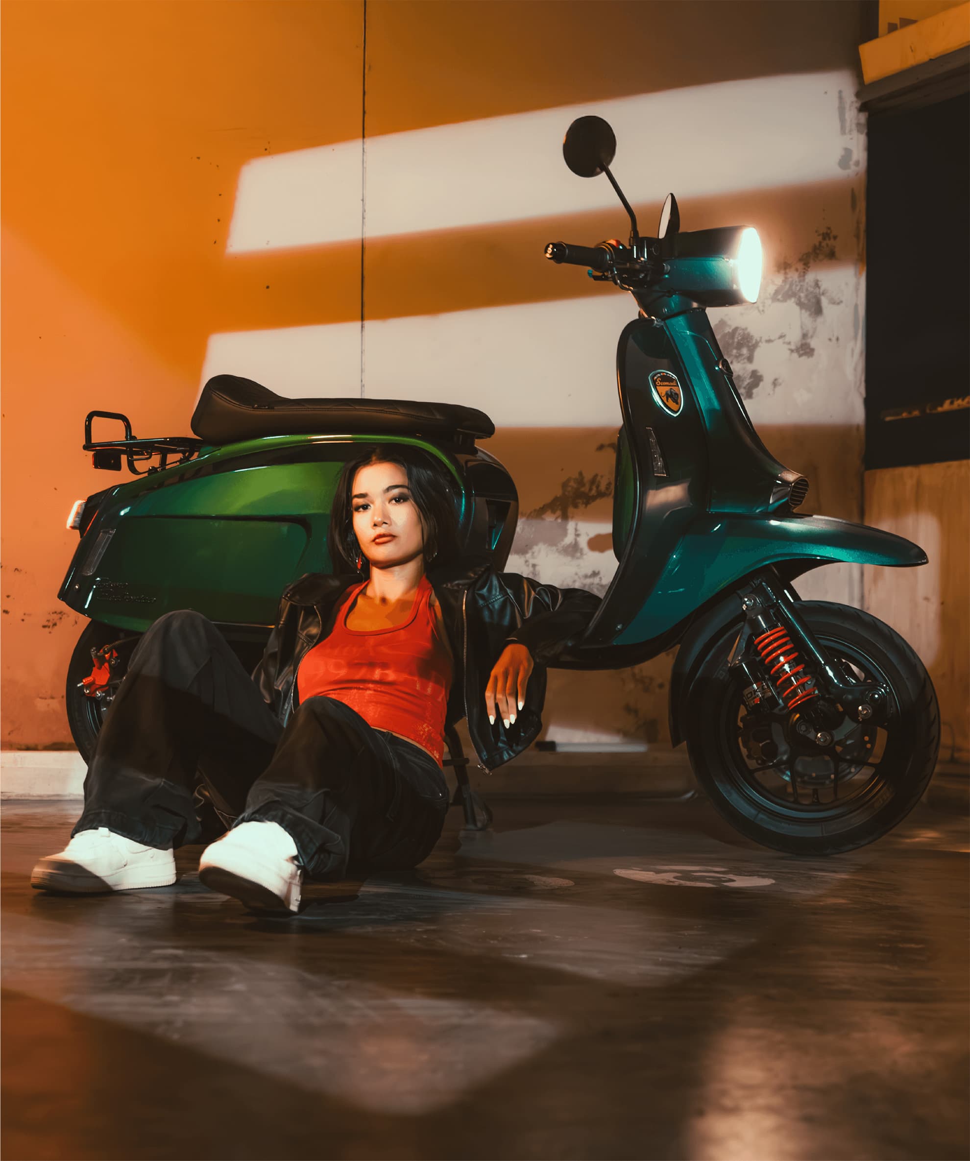 Rider sitting on the floor beside a teal Scomadi scooter in a garage