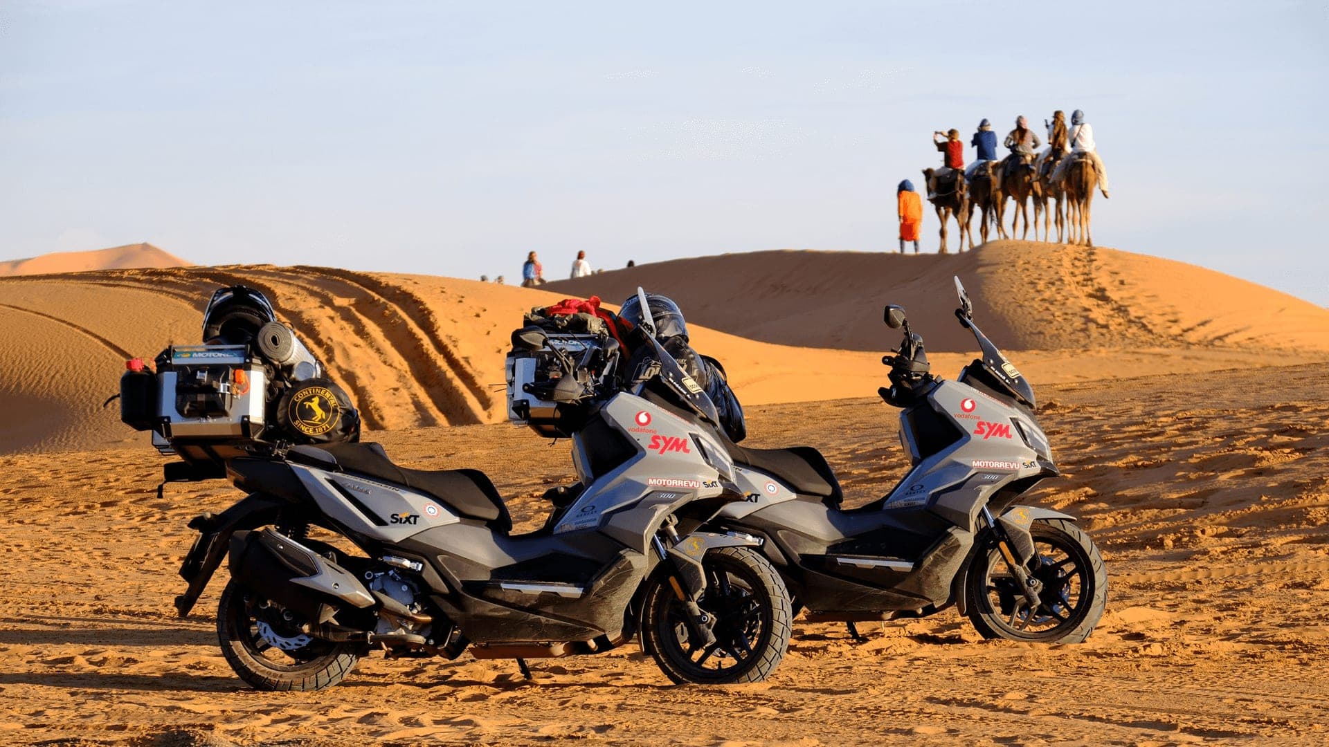 Two SYM ADX adventure scooters loaded for touring on a desert dune